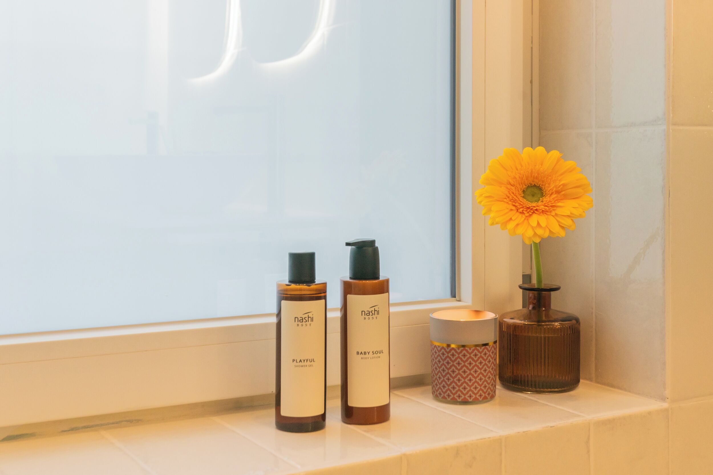 A bathroom shelf with two bottles of Nashi Baby Soul body lotion, a candle, and a vase with a yellow flower.