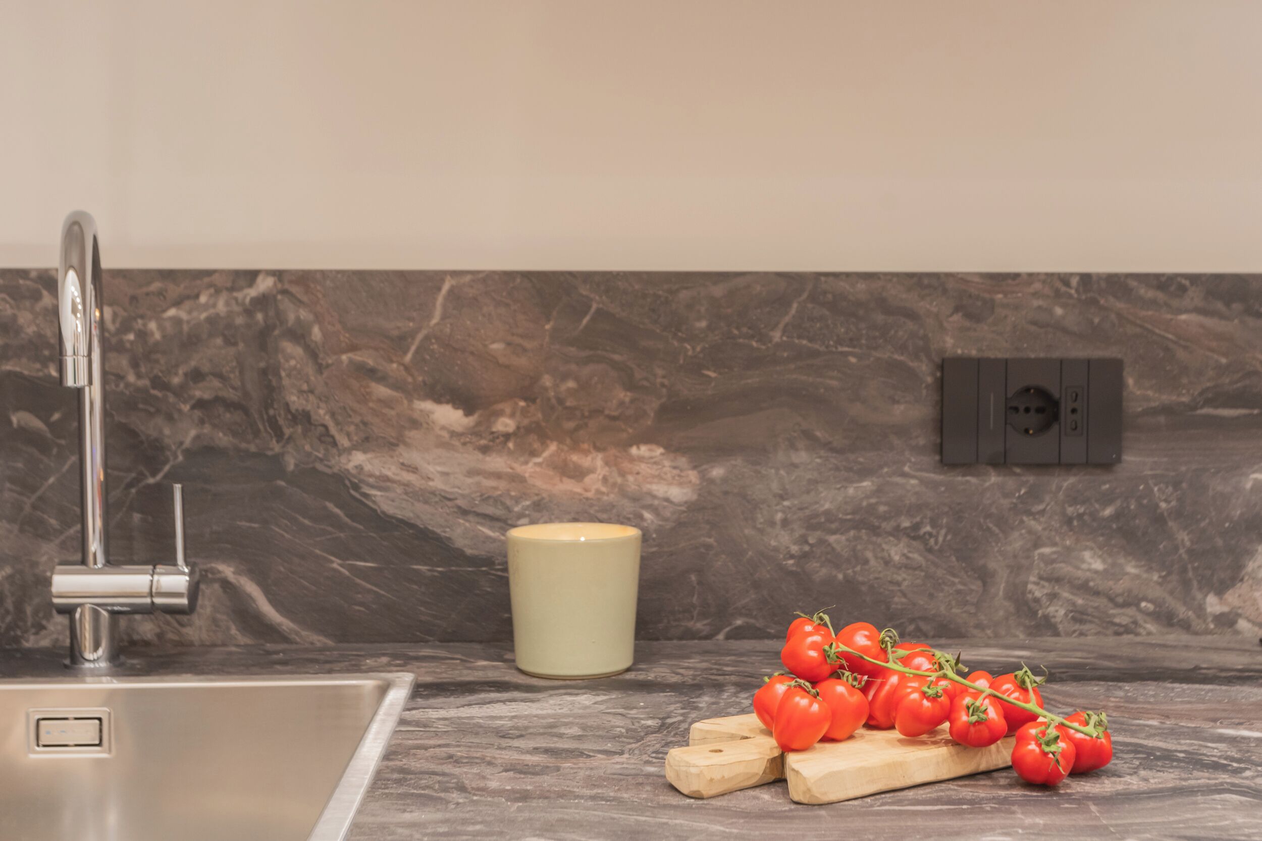 A kitchen with a stainless steel sink, a marble backsplash, and a wooden cutting board with tomatoes.