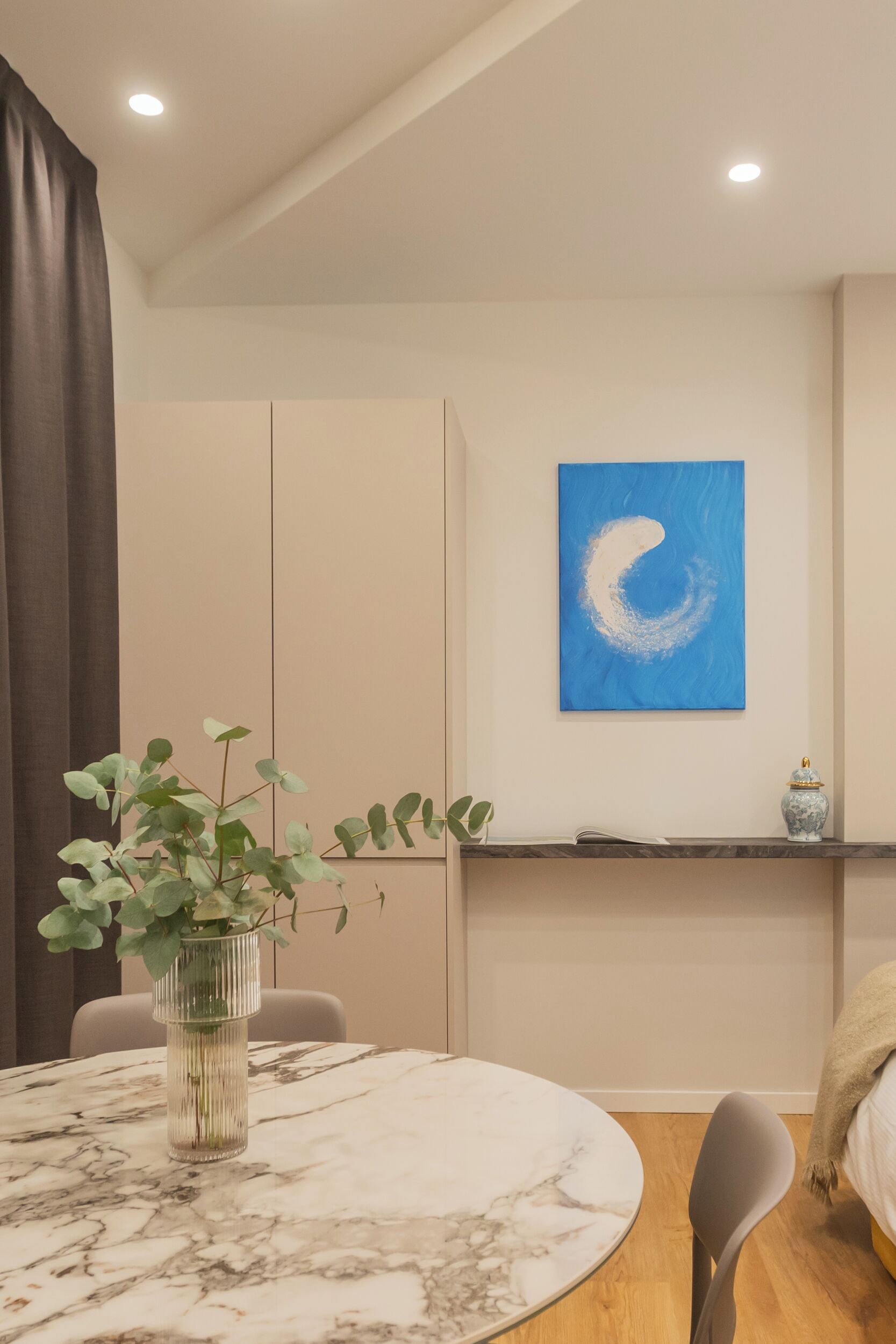 A modern room with a round marble table, a potted plant, a blue abstract painting, and a shelf with a decorative item.