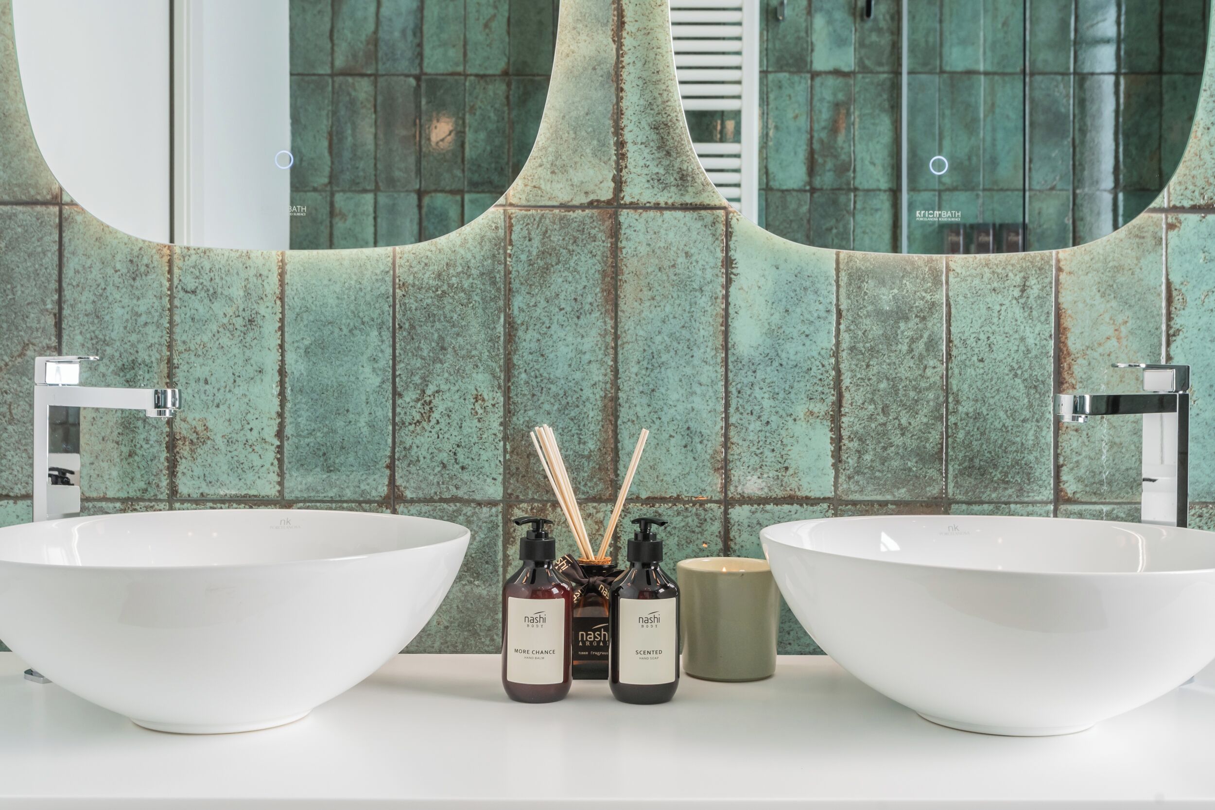 Une salle de bain avec deux lavabos blancs, un mur recouvert de carreaux verts et un miroir qui reflète la pièce.