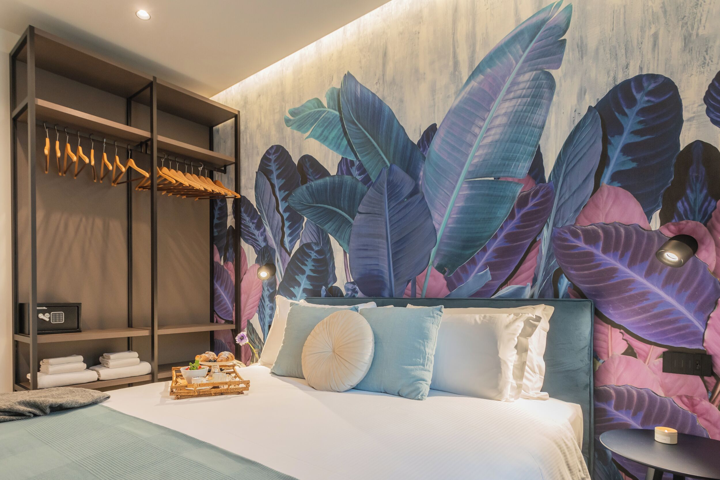 A bedroom with a large bed, a wardrobe with hanging clothes, and a wall mural of tropical leaves.