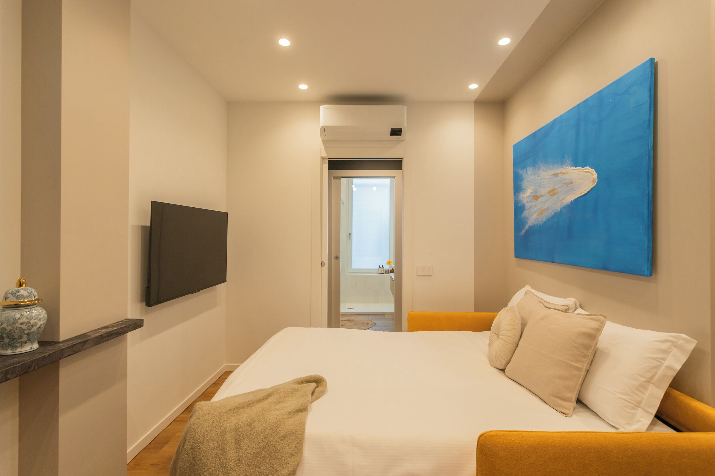 A hotel room with a bed, a flat-screen TV, a painting, and a bathroom visible through an open door.