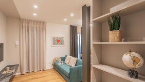 A modern living room with a teal sofa, wooden flooring, a bookshelf with decorative items, and a large window with curtains. - Rezia Apartments by Rent all Como (Como)