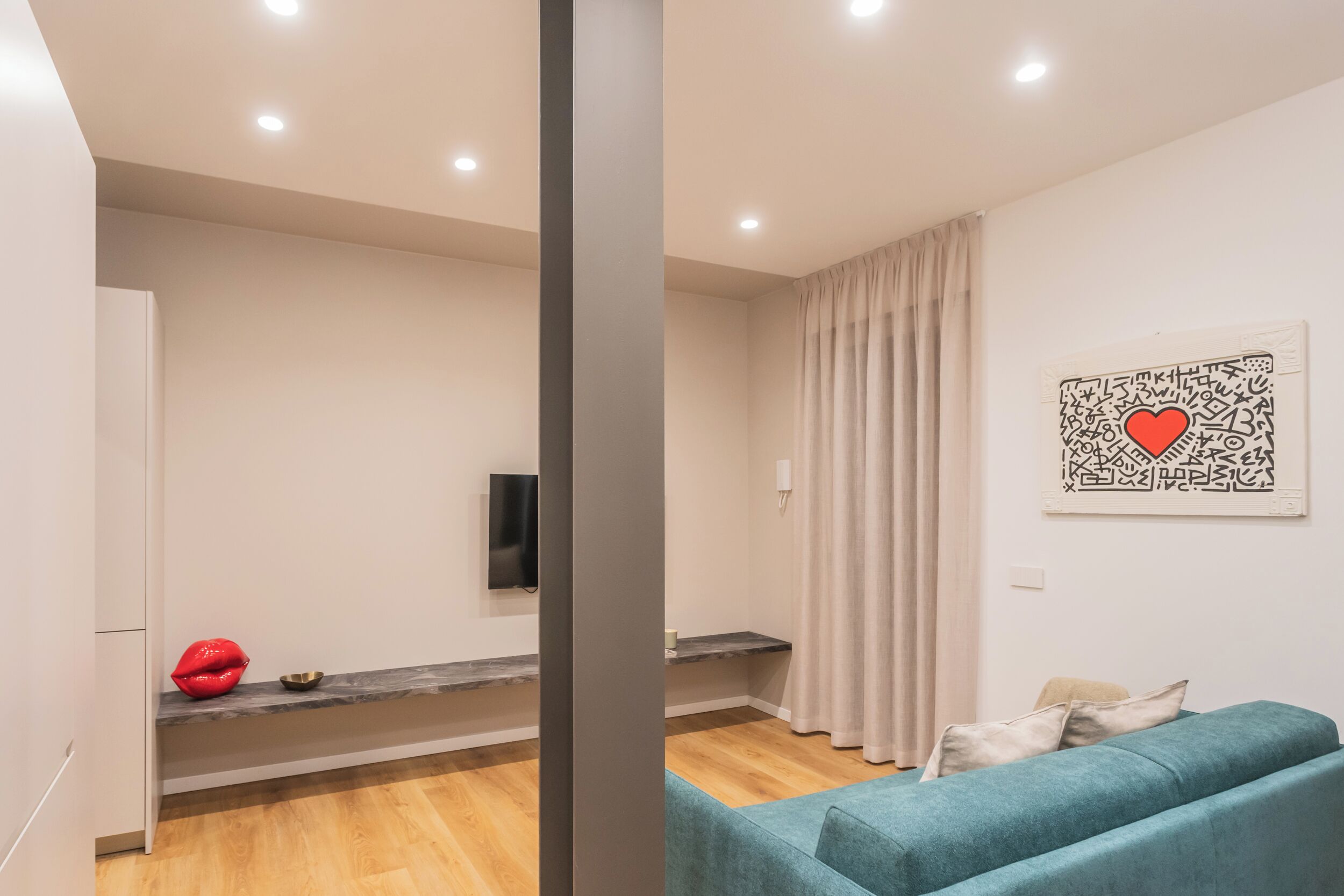 A modern living room with a teal sofa, a flat-screen TV mounted on the wall, a red decorative object, and a minimalist design.