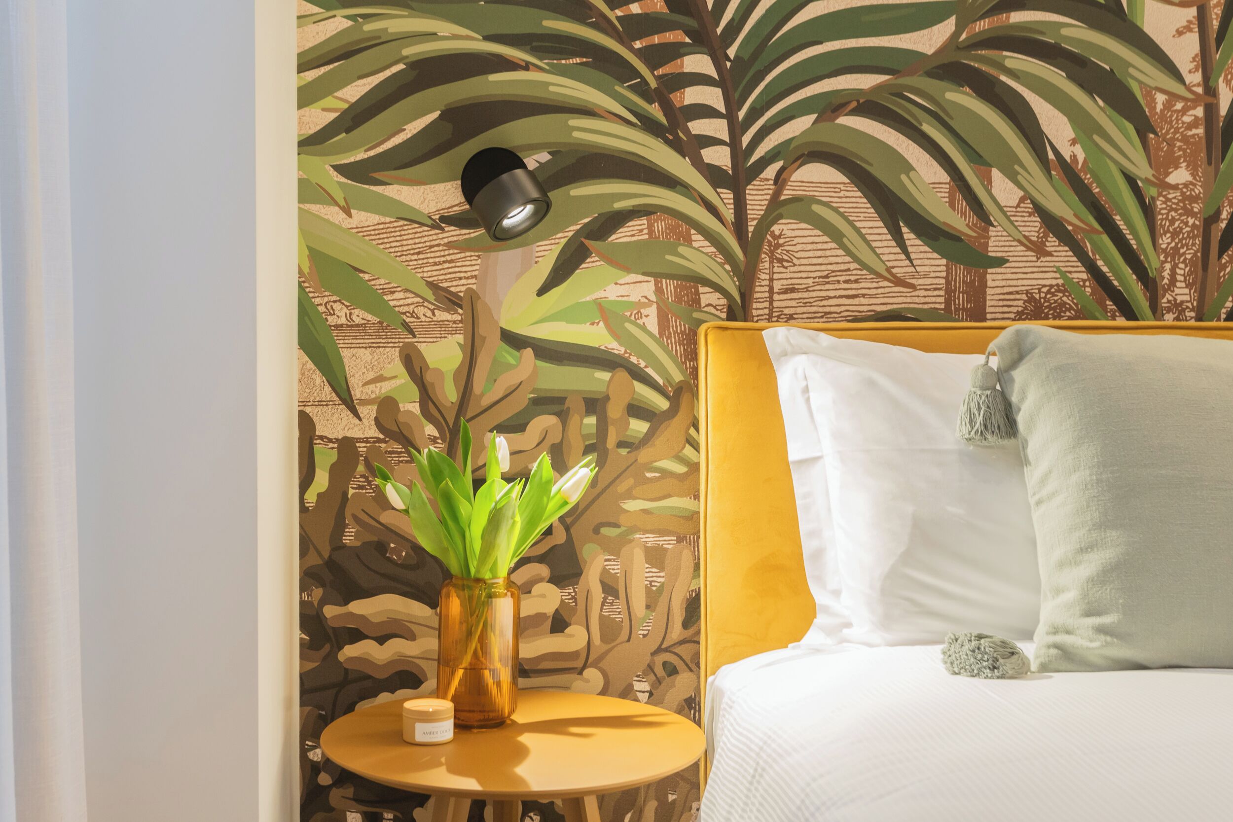 A room with a bed, a small round table with a plant and a candle, and a wall with a tropical mural.