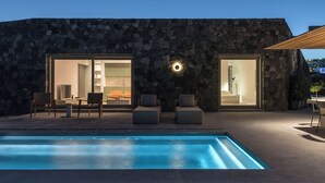 Un área de piscina moderna al aire libre con muros de piedra, un espacio de descanso con asientos y vistas al interior.