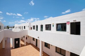 Un bâtiment blanc avec de nombreuses fenêtres et portes, une cour dallée et un ciel d’un bleu limpide.