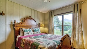 A bedroom with a wooden bed, a quilt, a window with curtains, and a view of greenery outside.