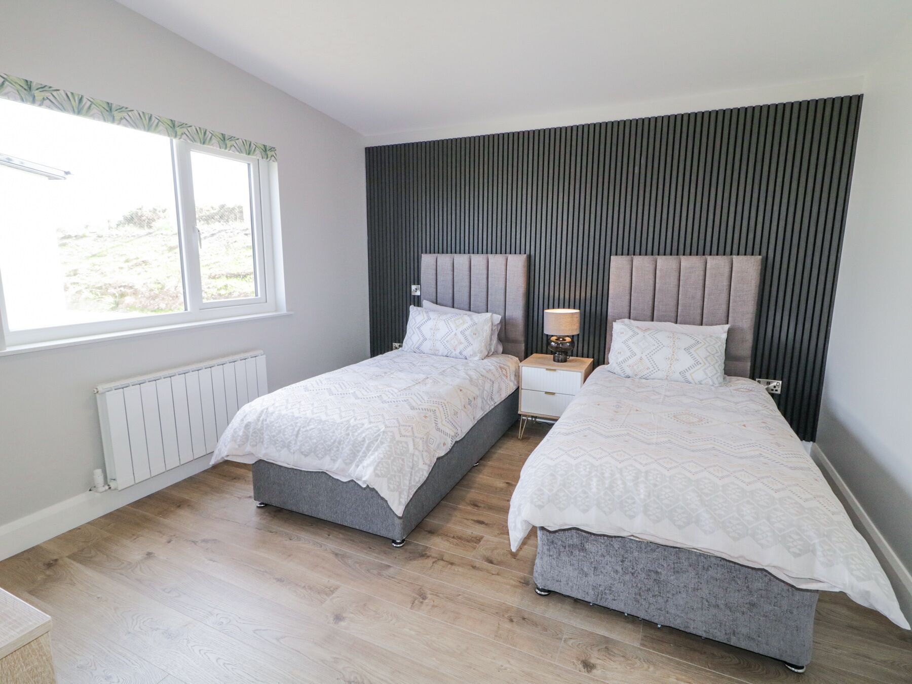 A bedroom with two beds, a window, a radiator, and a bedside table with a lamp.