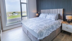 Une chambre avec une grande fenêtre, un lit recouvert d’une couvre-lit à motifs, une table de chevet avec une lampe, et un mur foncé orné de lignes verticales.