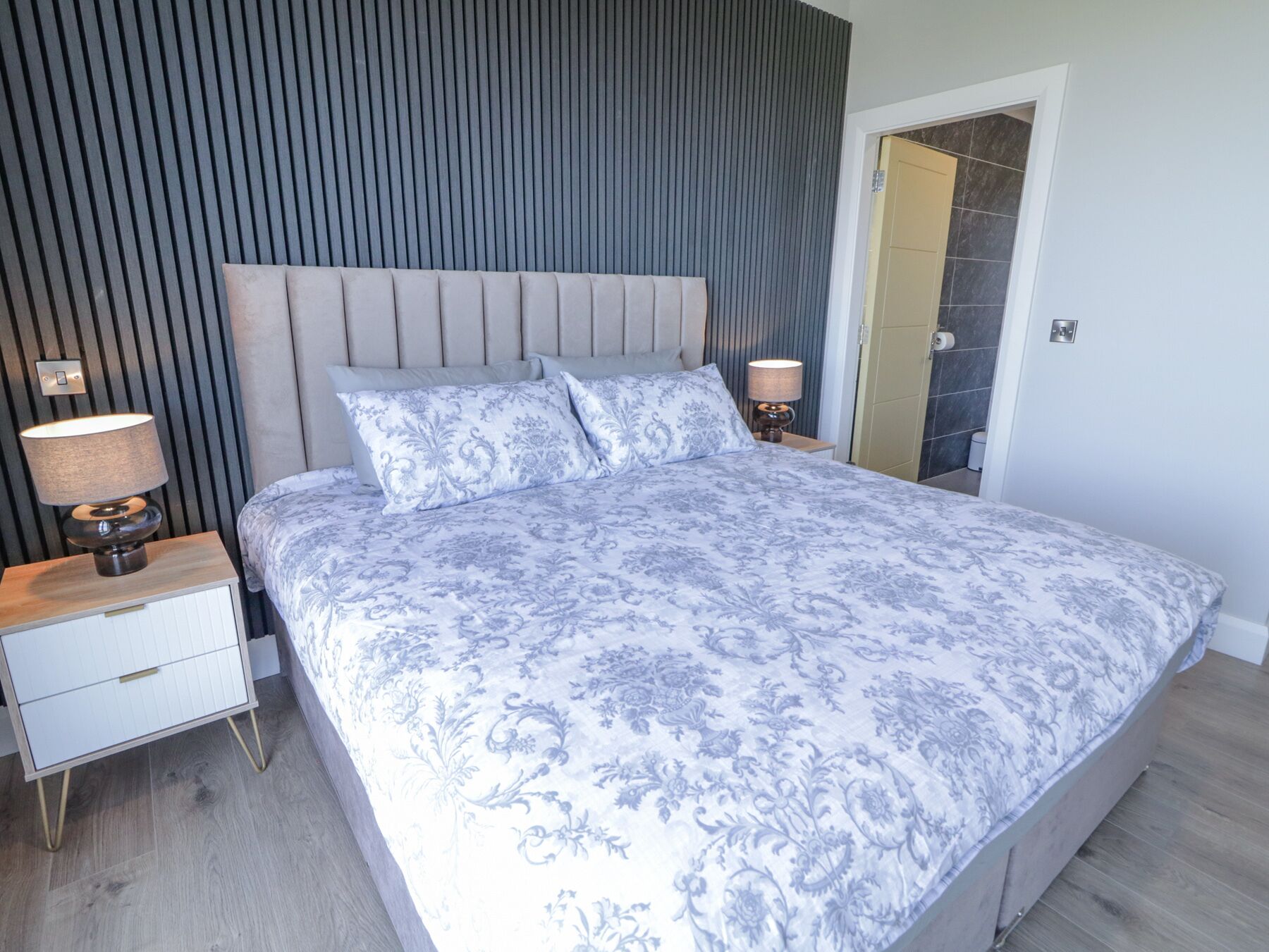 Un dormitorio con una cama grande, dos mesitas de noche con lámparas, un tocador de madera y una puerta que da a un baño.