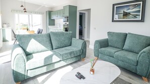 A modern living room with two green sofas, a coffee table, and a kitchen in the background. - Number 3 Villa - The View (Bunbeg)