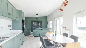 A modern kitchen with teal cabinets, a dining area with a round table and chairs, and a living area with a sofa and TV. - Number 3 Villa - The View (Bunbeg)
