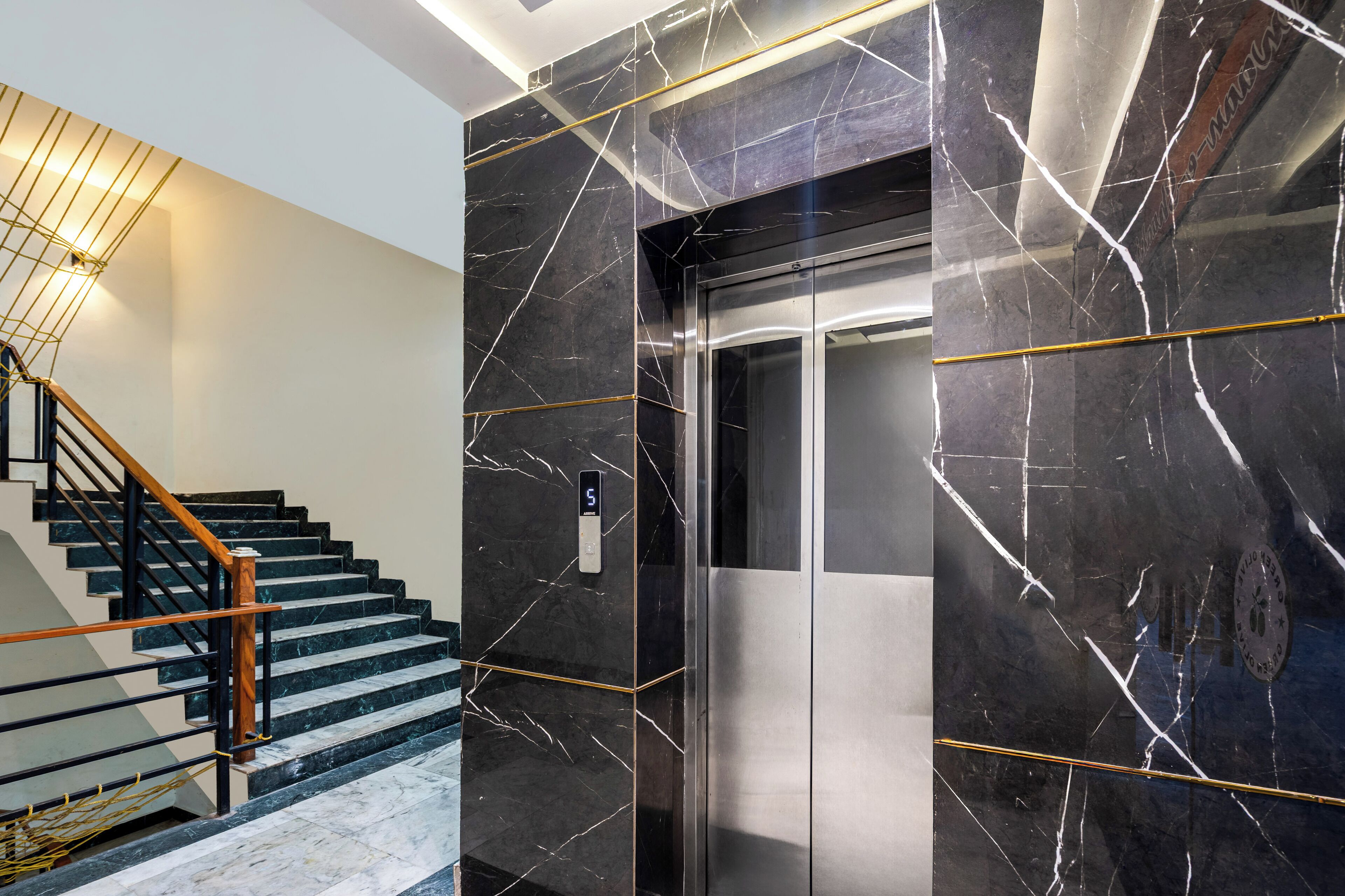 A modern elevator with a metallic finish, a marble wall with white veining, and a wooden staircase with metal railings.