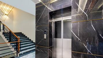 A modern elevator with a metallic finish, a marble wall with white veining, and a wooden staircase with metal railings.