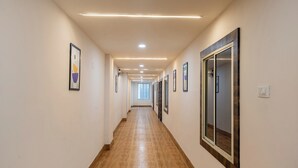 Um corredor longo e estreito, com piso de madeira e paredes brancas, com quadros emoldurados e um espelho grande em uma das laterais.