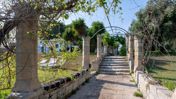 Un sendero de piedra que lleva a una estructura con arcos, rodeada de vegetación y un cielo despejado.