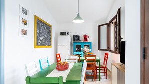 Un comedor con una mesa verde, sillas blancas, un bol rojo de fruta, un refrigerador blanco, un armario azul y una lámpara colgante.