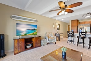 A living room with a large TV, a ceiling fan, a wooden cabinet, and a kitchen area with a refrigerator and bar stools. - Boutique-Inspired Tropical Décor (Kihei)