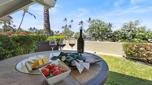 Ein Tisch mit Käseplatte, Erdbeeren, Weingläsern und einer Flasche, mit Blick auf einen Garten mit Palmen und Blumen.