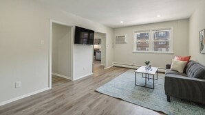 A spacious living room with hardwood flooring, a grey sofa, a coffee table, and a large window. - Pine Brook 618 I Cozy & Roomy I Gym (Newark)