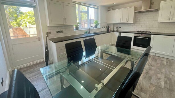 A modern kitchen with white cabinets, a glass dining table, black chairs, a gas stove, and a window with a view of greenery. - Spacious 4-bedroom House - Garden & Free Parking (Faversham)