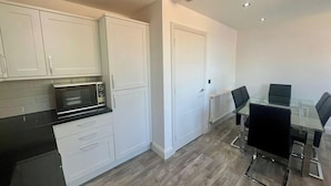 A modern kitchen with white cabinets, a black countertop, and a built-in microwave. There is a glass dining table with black chairs and a white door in the background. - Spacious 4-bedroom House - Garden & Free Parking (Faversham)