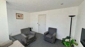 A living room with two grey armchairs, a beige sofa, a white door, a framed picture on the wall, and a floor lamp. - Spacious 4-bedroom House - Garden & Free Parking (Faversham)