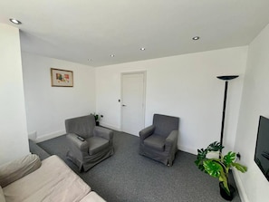 Una sala de estar con dos sillones grises, un sofá beige, una puerta blanca, un cuadro enmarcado en la pared y una lámpara de pie.