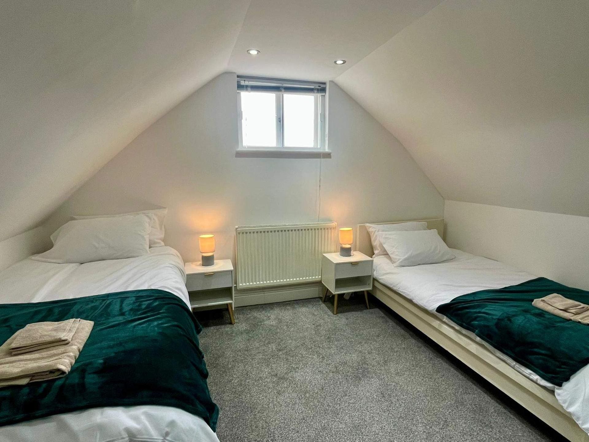 Un dormitorio estilo altillo, pequeño y triangular, con dos camas, cada una con una mesita de noche y lámpara, y un radiador en medio.
