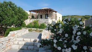 Un edificio con pareti in pietra, una pergola in legno, fiori bianchi e un cielo azzurro limpido.