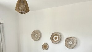 Ein ordentlich bezogenes Bett mit weißen Leinen, einer beige farbenen Tagesdecke und einem gefalteten Handtuch. Darüber an der Wand hängen drei gewebte Kunstwerke.