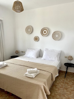 A neatly made bed with white linens, a beige throw blanket, and a folded towel on the bed. There are three wall-mounted woven art pieces above the bed.