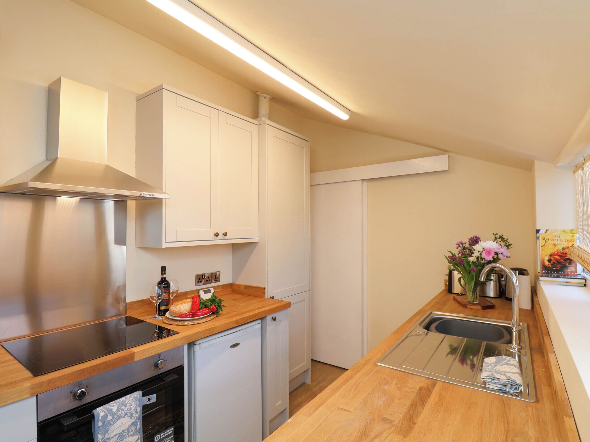Cocina compacta con horno integrado, un refrigerador pequeño, armarios blancos, una campana extractora de acero inoxidable y una encimera de madera.