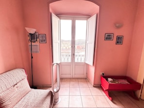 Un petit balcon donnant sur des immeubles, une porte blanche, un banc rouge et un lampadaire.