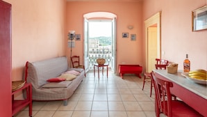 Ein gemütliches Zimmer mit einer Couch, Sesseln, einem Esstisch mit einer Obstschüssel und Blick durch eine offene Tür.