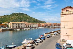 Une rue bordée de bâtiments avec des balcons, donnant sur le front de mer, où sont garées des voitures et où des bateaux sont amarrés le long de la rivière.