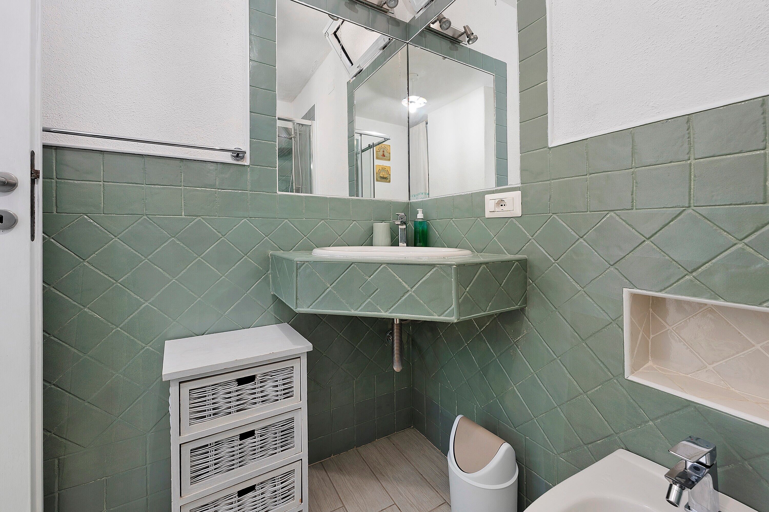 Une salle de bain avec un lavabo blanc, un miroir et des murs recouverts de carreaux verts.