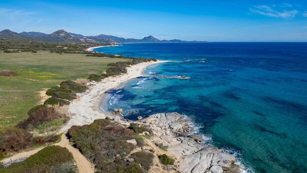 Un paysage côtier avec une plage, une eau d’un bleu limpide et des montagnes à l’horizon.