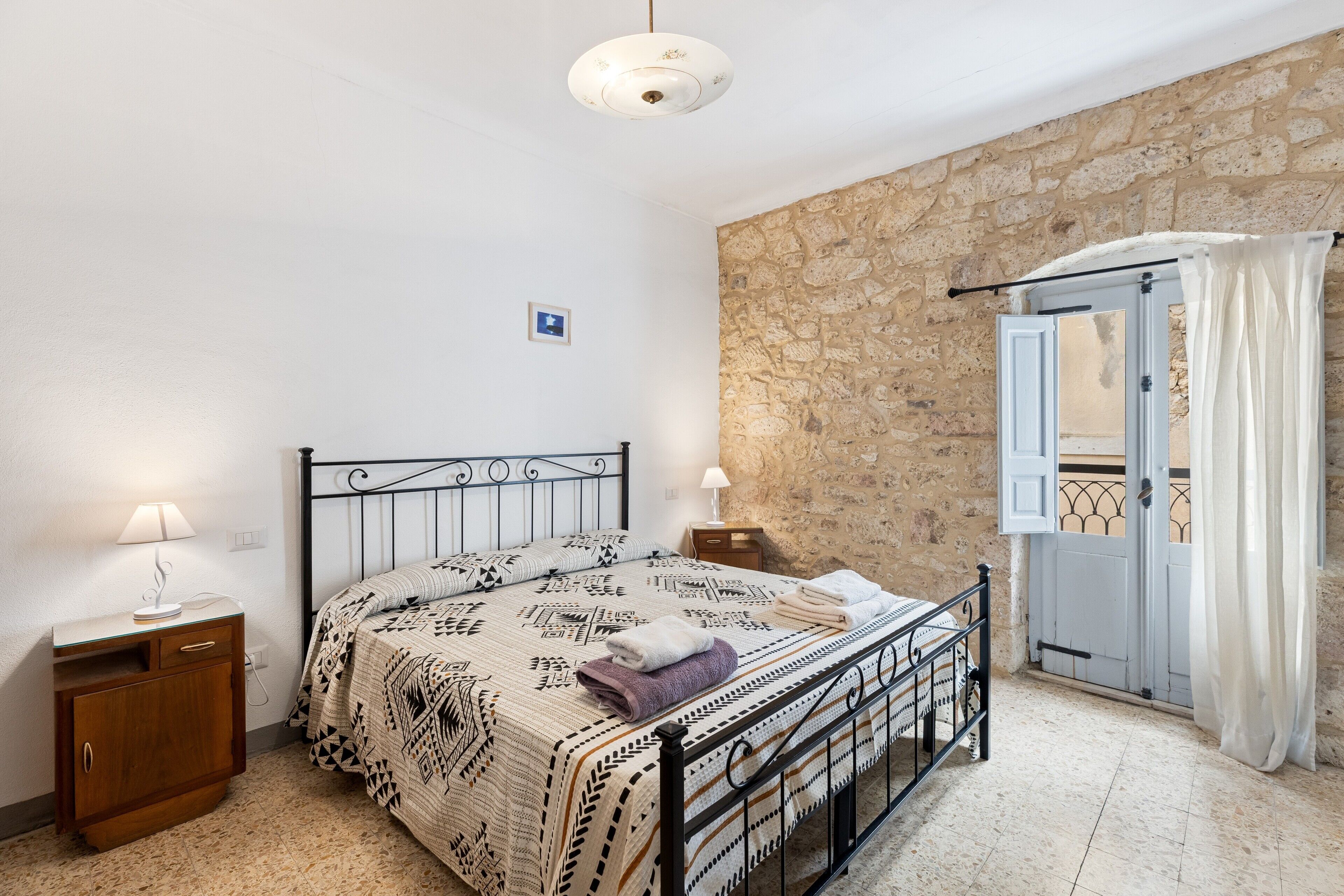 A bedroom with a bed, bedside tables, a stone wall, a door with a balcony, and a ceiling light.