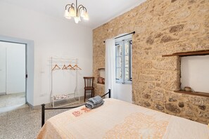A bedroom with a stone wall, a bed with a patterned bedspread, a hanging chair, a wooden chair, a window with curtains, and a chandelier. - Holiday Home 'Domo Mea - La Tua Casa In Sardegna' with Terrace, Balcony and Wi-Fi (Cuglieri)