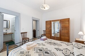 Une chambre à coucher avec un lit, une armoire en bois, une chaise, une petite table avec une lampe, et une salle de bain visible à travers une porte ouverte.