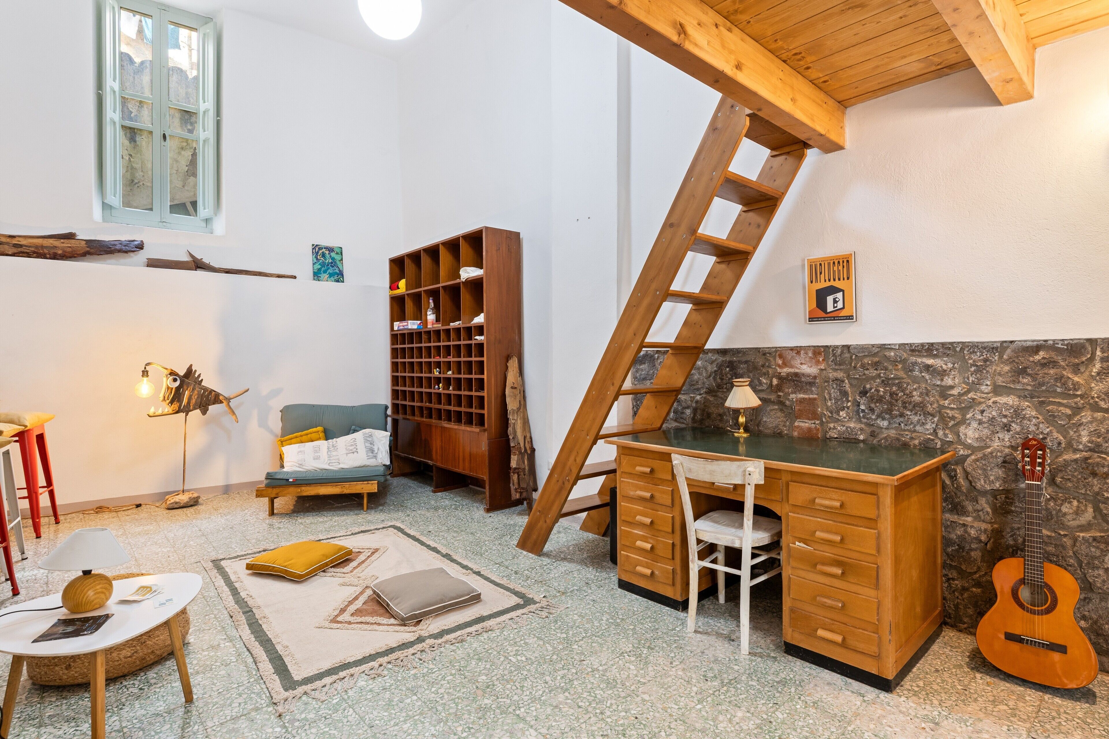 Une pièce avec un bureau en bois, une guitare, une bibliothèque, une échelle et un mur en pierre.