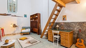 Une pièce avec un bureau en bois, une guitare, une bibliothèque, une échelle et un mur en pierre.