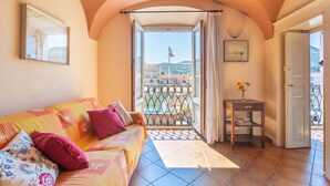 Ein gemütliches Zimmer mit Balkon, einer Couch mit Kissen, einem kleinen Tisch mit einer Vase und einer Tür nach draußen.