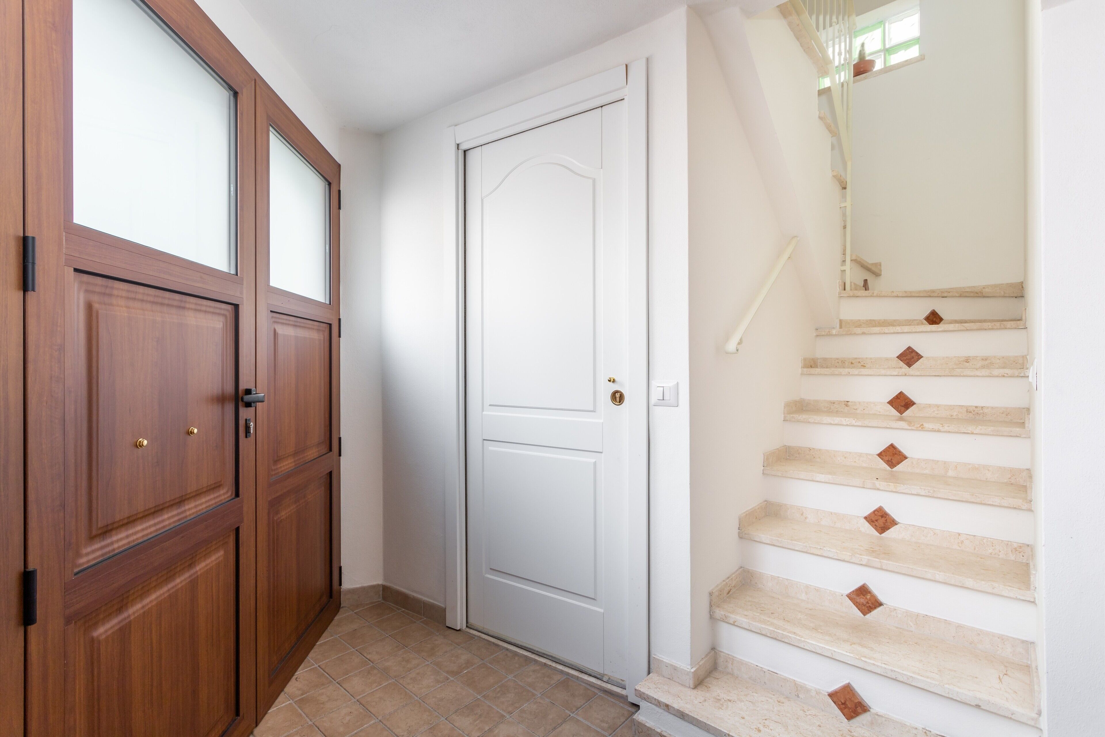 Un pasillo con una puerta de madera, una puerta blanca y una escalera con peldaños color beige y detalles en marrón.