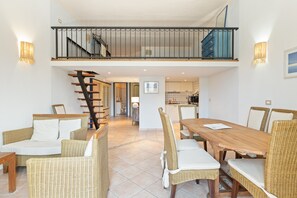 A spacious dining area with wicker furniture, a staircase leading to a balcony, and a kitchen in the background.