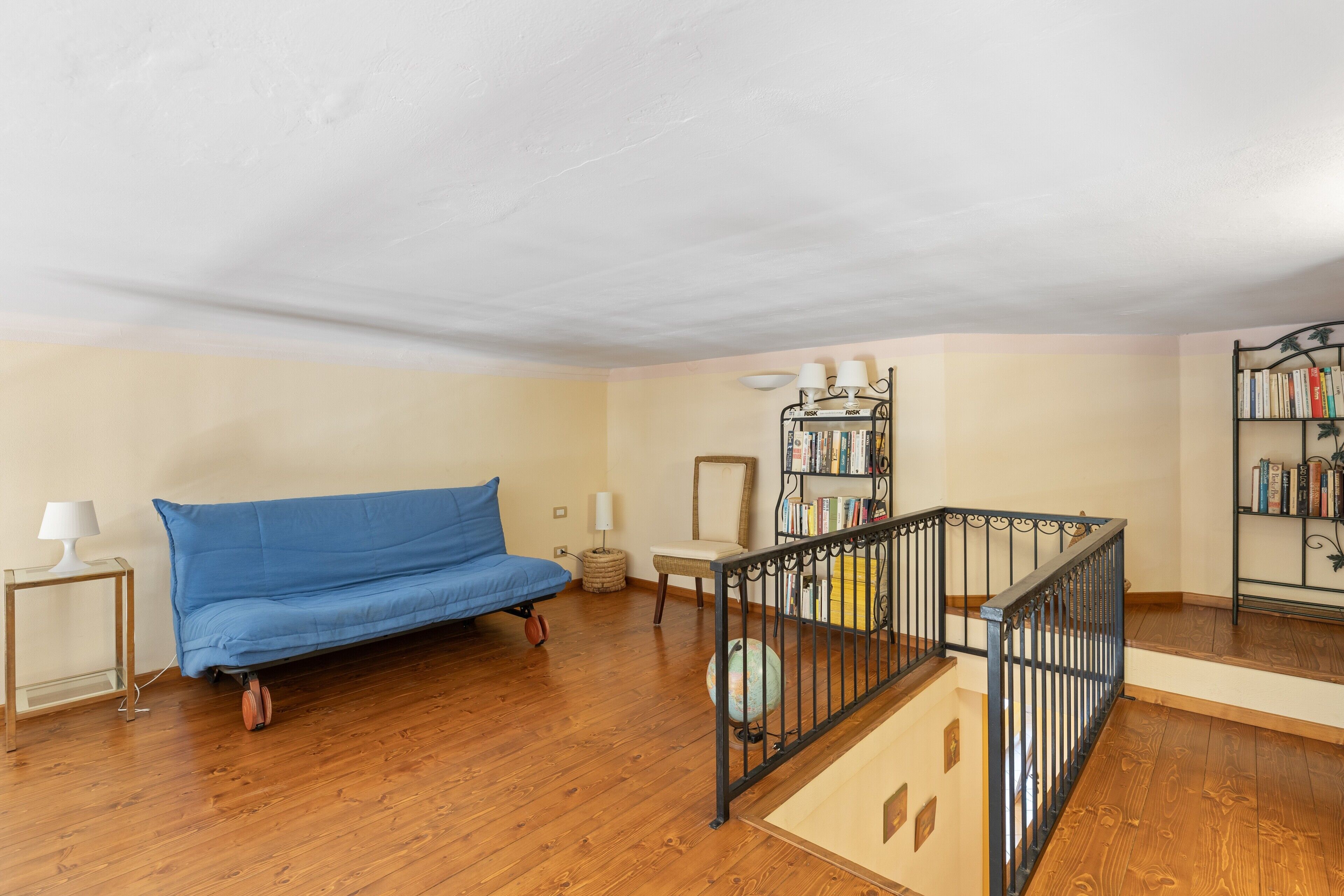 Ein Zimmer mit einem blauen Sofa, einem kleinen Tisch mit Lampe, einem Bücherregal und einer Treppe mit Metallgeländer.