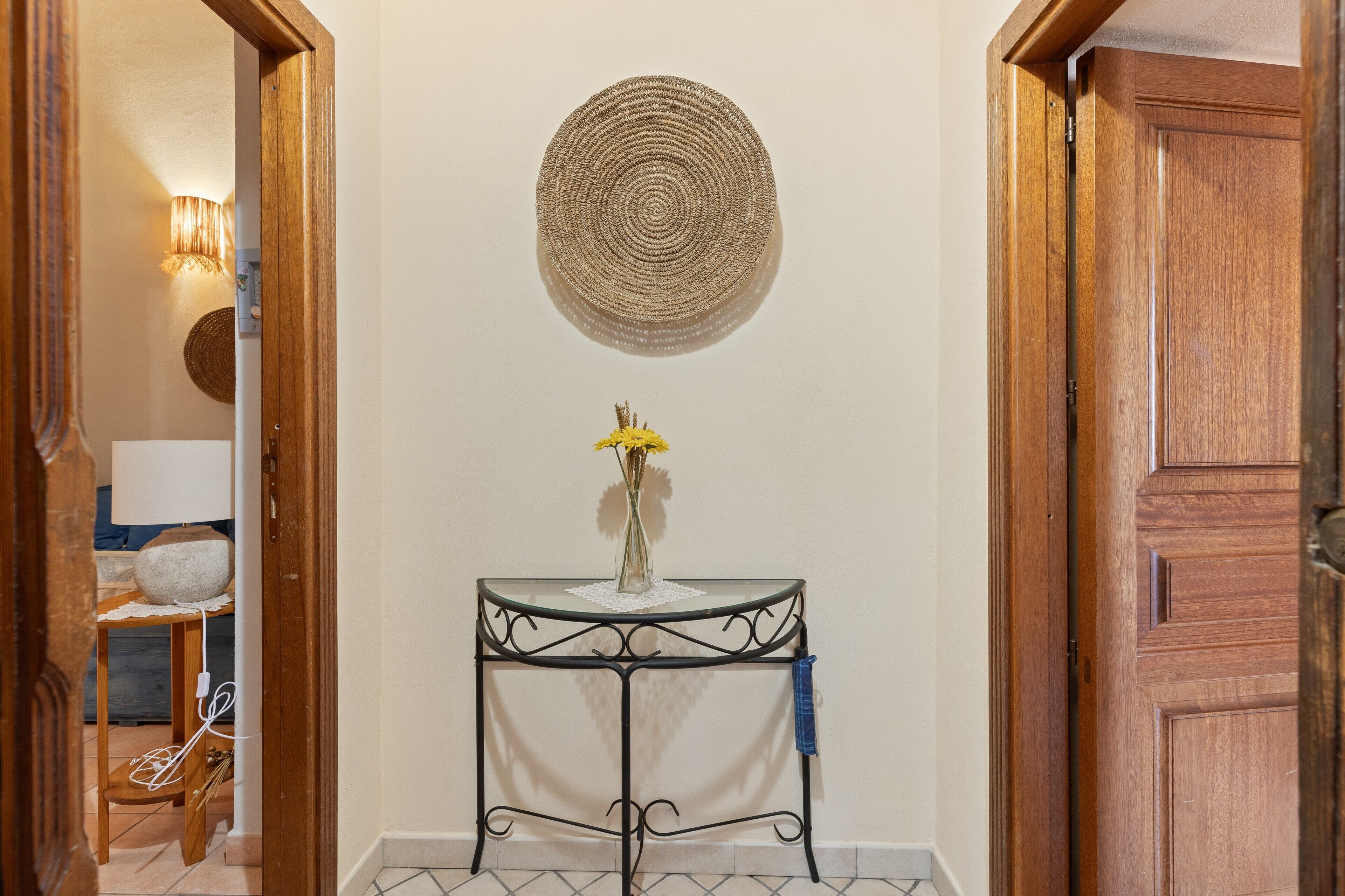 Un couloir orné d’une tapisserie murale ronde en fibres tressées, une petite table sur laquelle est posé un vase rempli de fleurs jaunes, et une porte en bois.