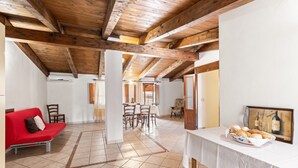A spacious room with a tiled floor, wooden ceiling beams, a dining area with chairs and a table, a red sofa, and a table with bread and a bottle.